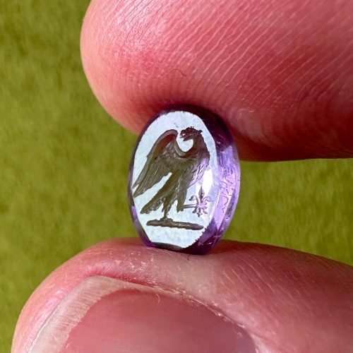 Small amethyst intaglio with facet back with imagery of an eagle rising, wings addorsed in preparation for flight, with a fleur-de-lis in its talons